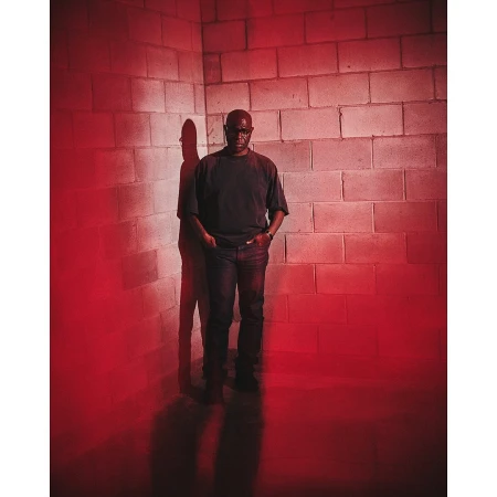 Portrait of a Black man standing in front of a cinder block wall, cast in dramatic red lighting with a noticeable shadow. He wears casual clothes.