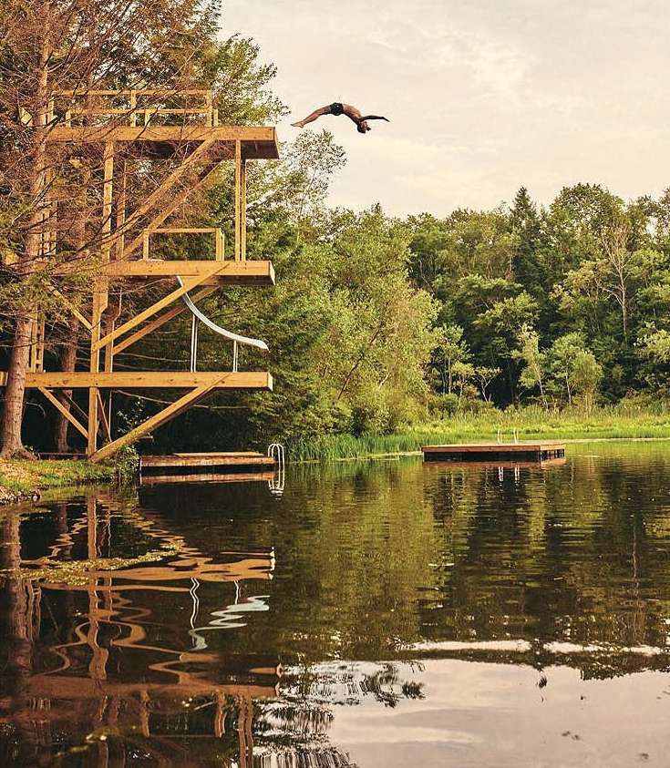 Person dives off a wooden platform into a lake surrounded by lush green trees under a bright sky in a daylight outdoor scene.