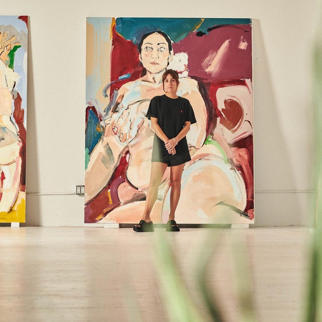 Woman stands in front of large painted portrait in a studio. The artwork features a stylized female figure with bold, expressive brushstrokes.