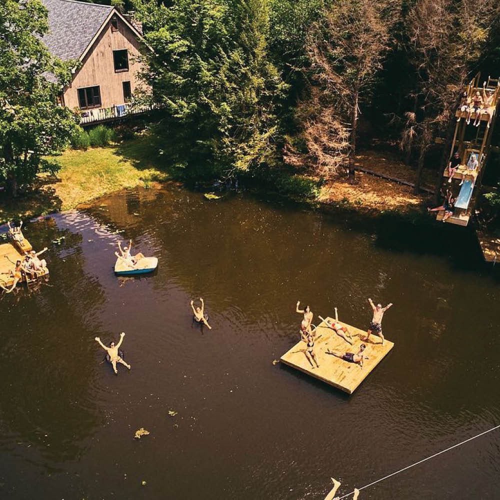 Aerial view of group enjoying summer on a lake with a house and trees in the background; people swim, float, and relax on rafts.