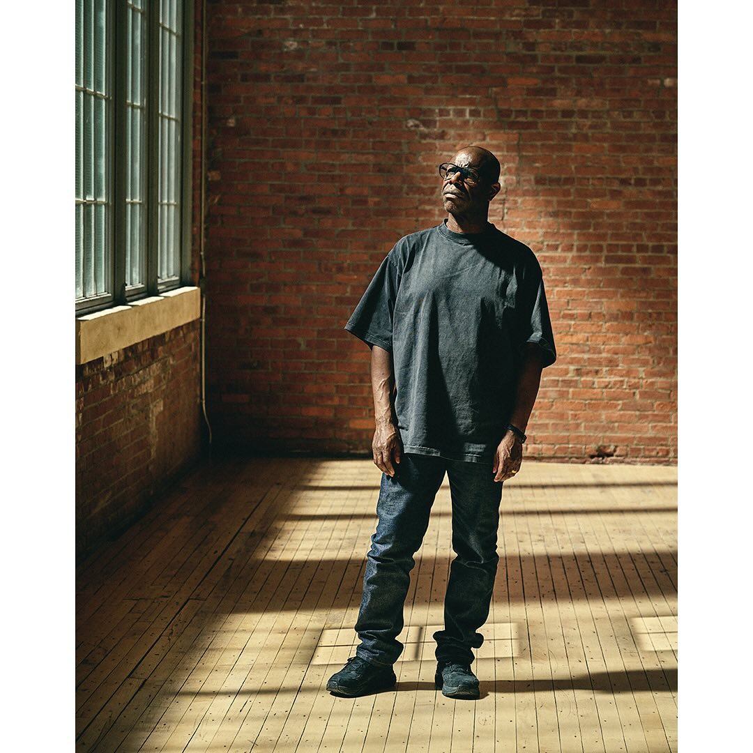 Adult man standing indoors near a window and a brick wall wearing a grey t-shirt, jeans, and sneakers looking off into the distance.