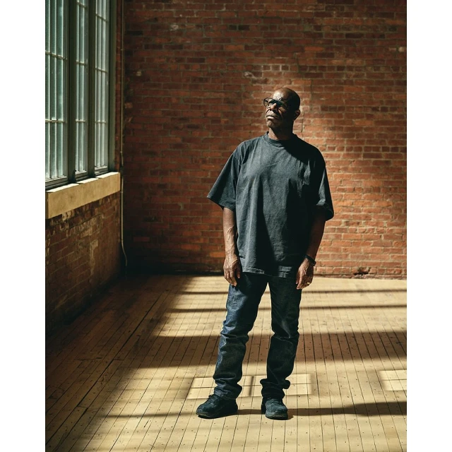 Adult man standing indoors near a window and a brick wall wearing a grey t-shirt, jeans, and sneakers looking off into the distance.
