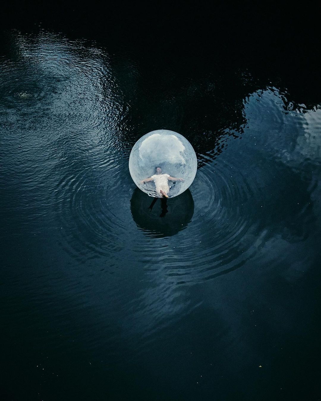 Aerial shot of a shirtless man floating in a clear, inflatable bubble on a dark lake surrounded by ripples on a cloudy day.