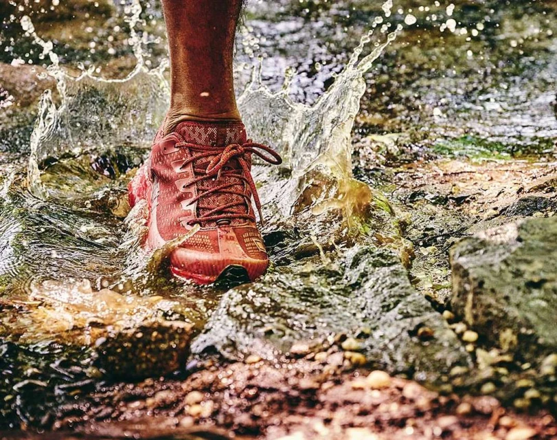 Close-up of a person's foot stepping into water with a red trail running shoe, creating a splash. Low angle shows texture of rocks.