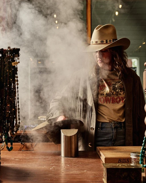 Woman in cowboy hat and fringe jacket stands behind wooden table with beaded necklaces and smoke rising from a brass container.