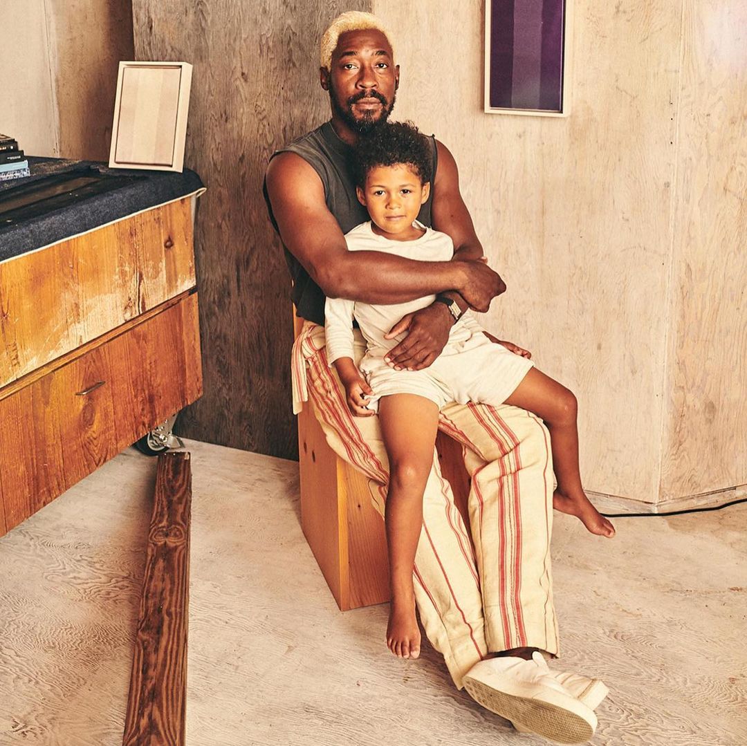 An African-American man with bleached hair embraces a young boy seated on his lap, set against a neutral backdrop with minimalist decor.