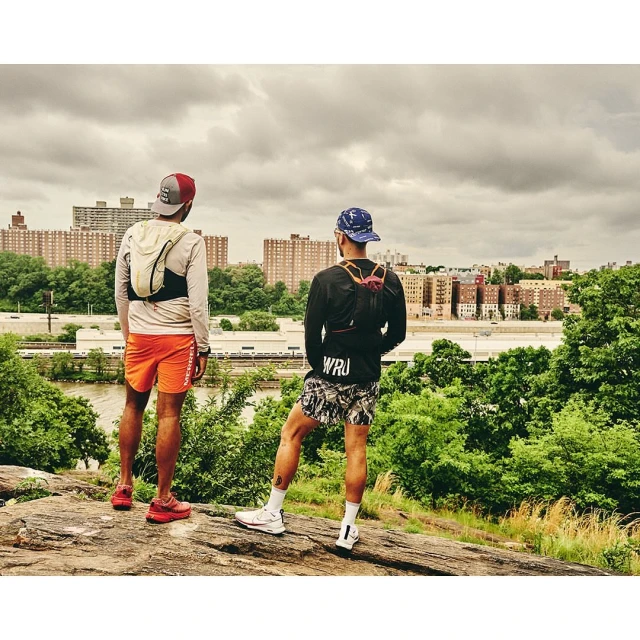Two adult males in athletic wear stand on a rock outcropping overlooking a city skyline and river, showcasing outdoor fitness and urban nature.