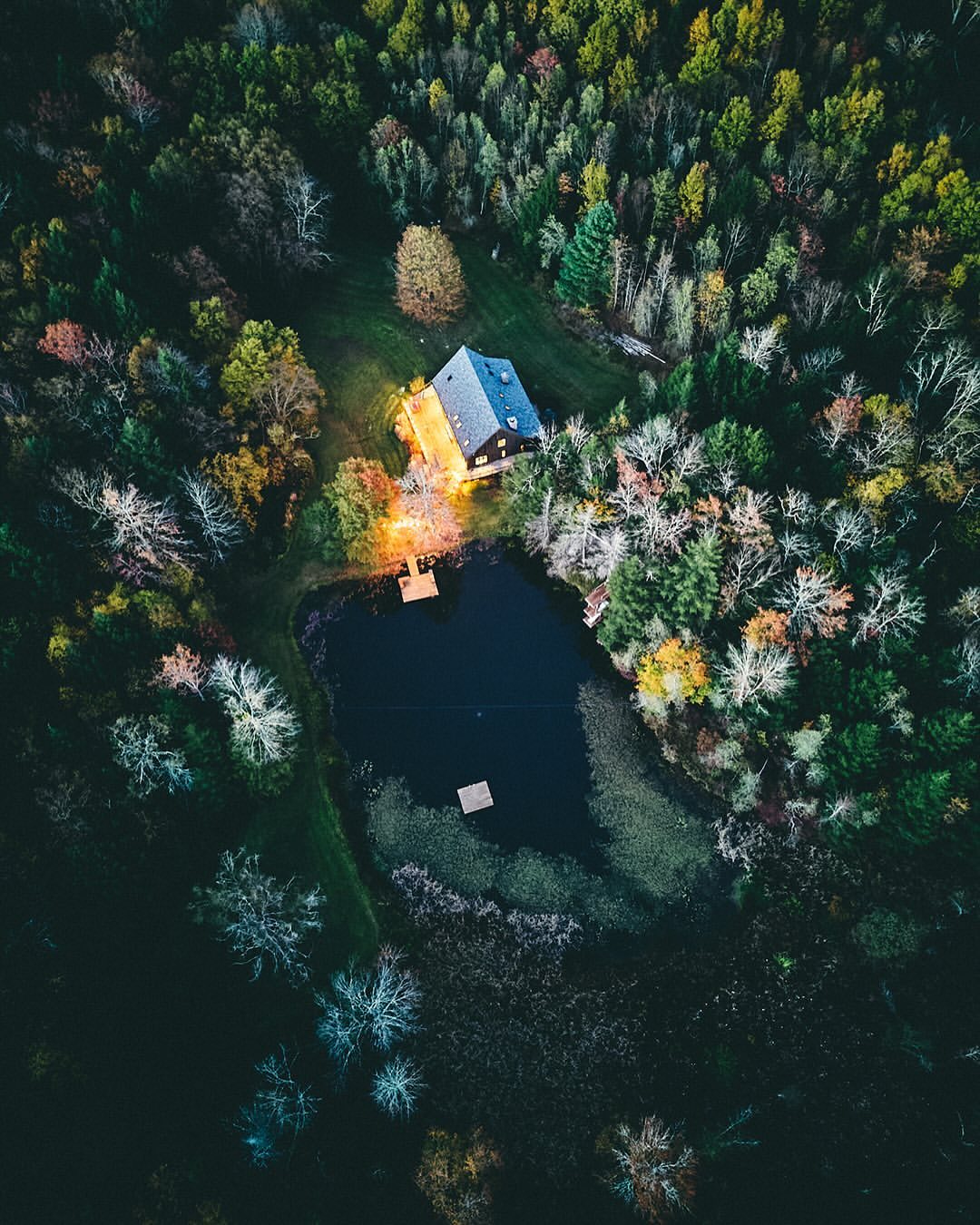 Aerial view of a dark lake surrounded by a lush forest and a small wooden house illuminated with warm light. Nature scene.