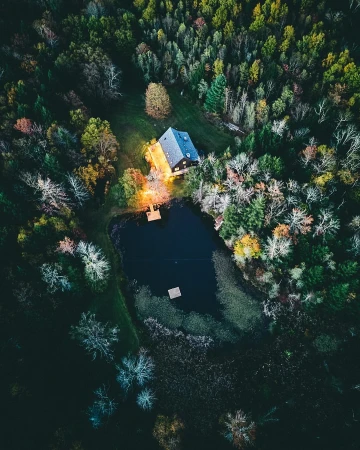 Aerial view of a dark lake surrounded by a lush forest and a small wooden house illuminated with warm light. Nature scene.