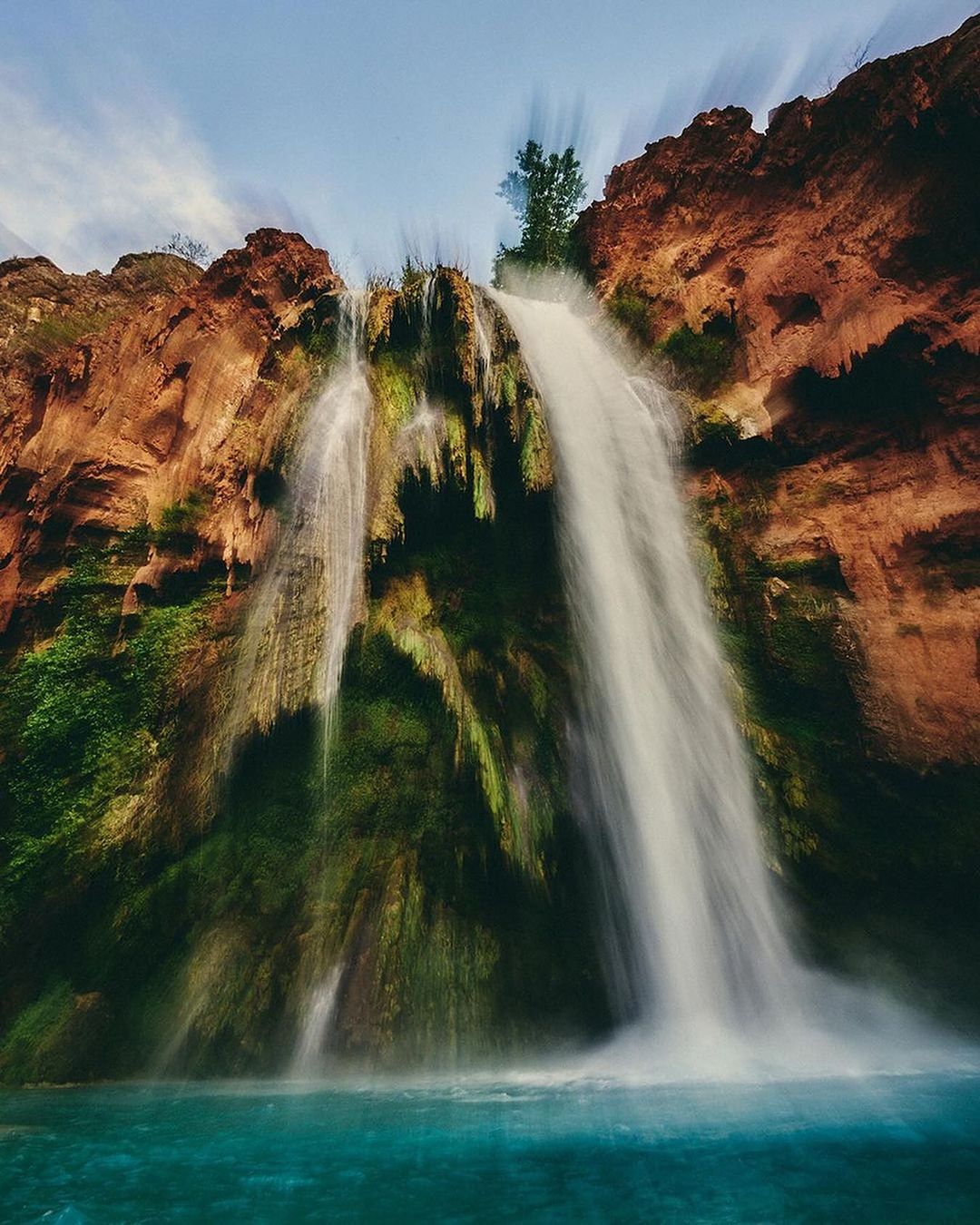 Scenic view of a majestic waterfall cascading into a clear turquoise pool, surrounded by reddish-brown rock formations and lush green foliage.