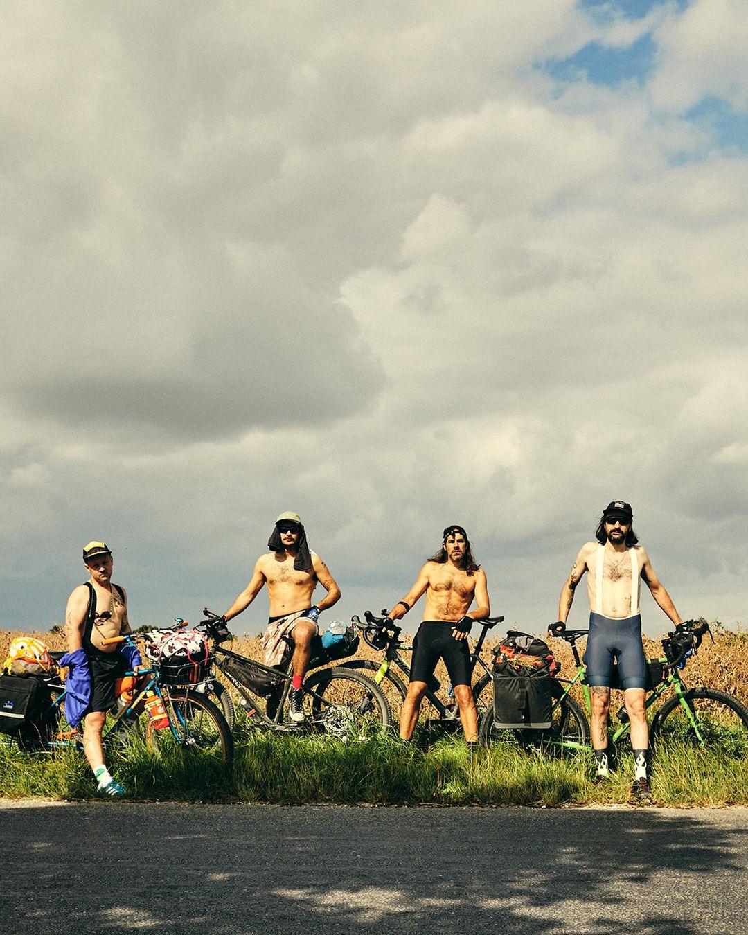 Four cyclists pose with their touring bikes on a rural road under a cloudy sky. They are shirtless, wearing hats and cycling shorts.