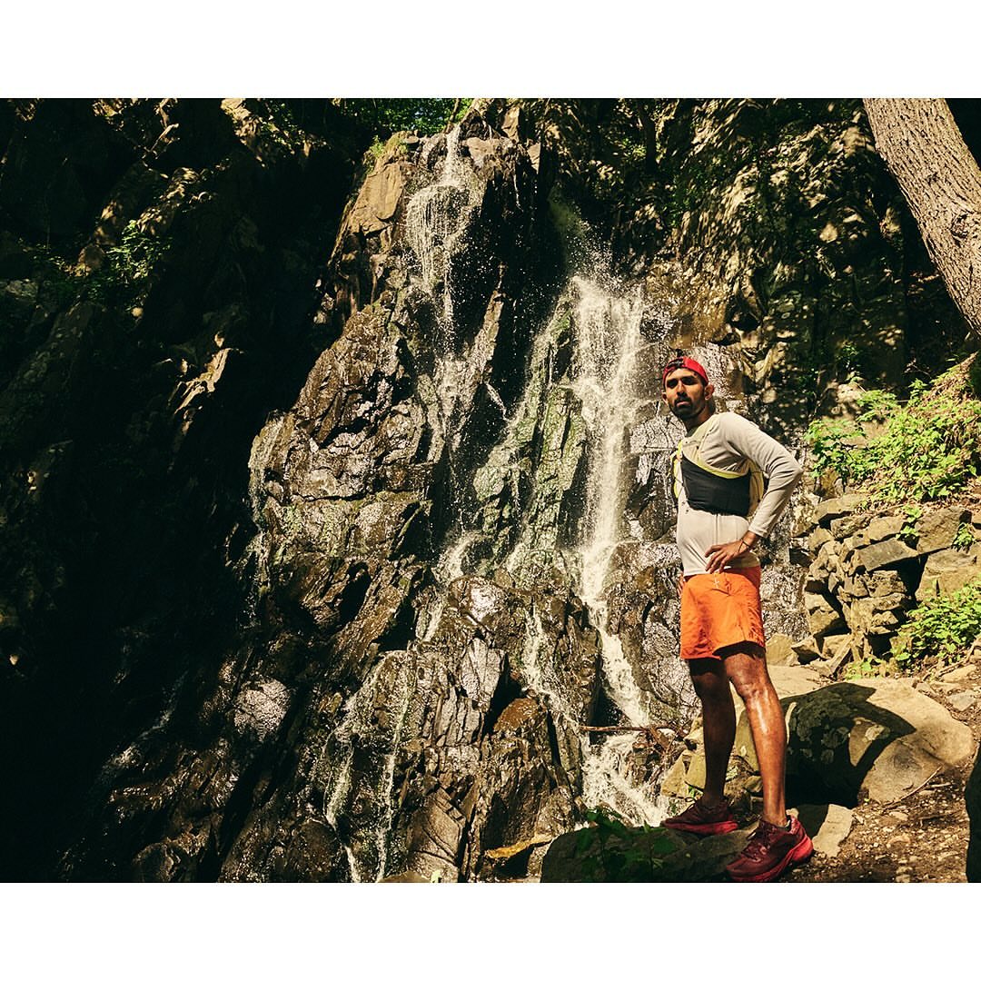 Adult male stands near a scenic waterfall wearing athletic clothes and a hydration pack in a forest setting on a sunny day.