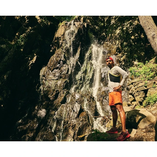 Adult male stands near a scenic waterfall wearing athletic clothes and a hydration pack in a forest setting on a sunny day.