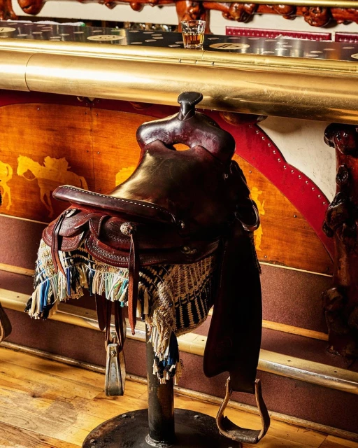 Leather western saddle on a stand in an indoor setting with a liquor glass in the background, creating a rustic bar scene.