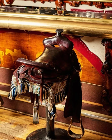 Leather western saddle on a stand in an indoor setting with a liquor glass in the background, creating a rustic bar scene.
