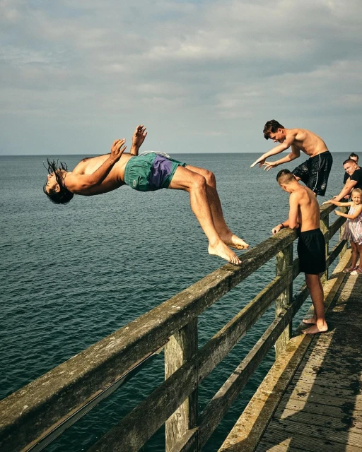 Man doing a backflip off a wooden pier with friends, overlooking the ocean on a sunny day with cloudy skies in the background.