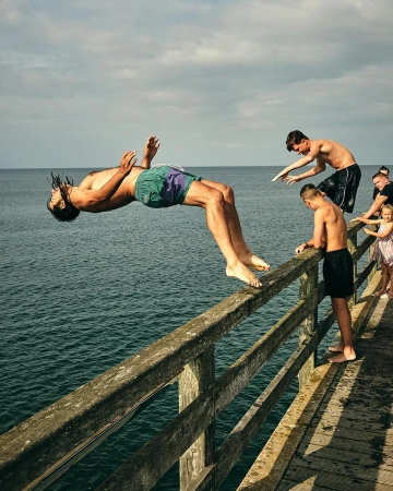 Man doing a backflip off a wooden pier with friends, overlooking the ocean on a sunny day with cloudy skies in the background.