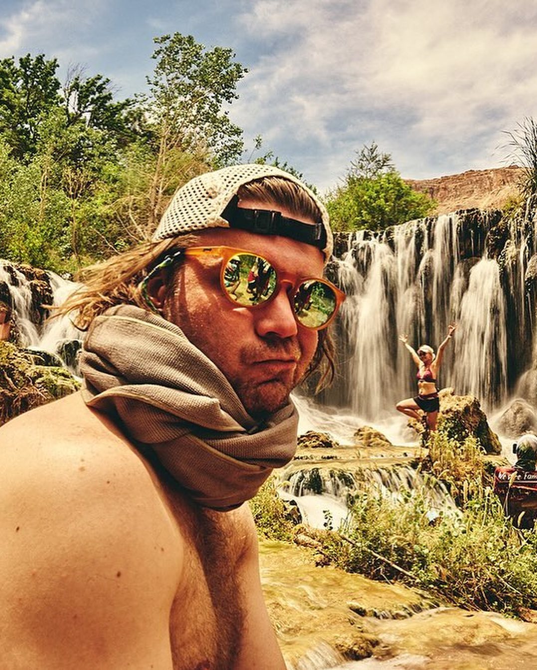 Man wearing a white knit cap and amber sunglasses with a heavy scarf around his neck near a rushing waterfall.