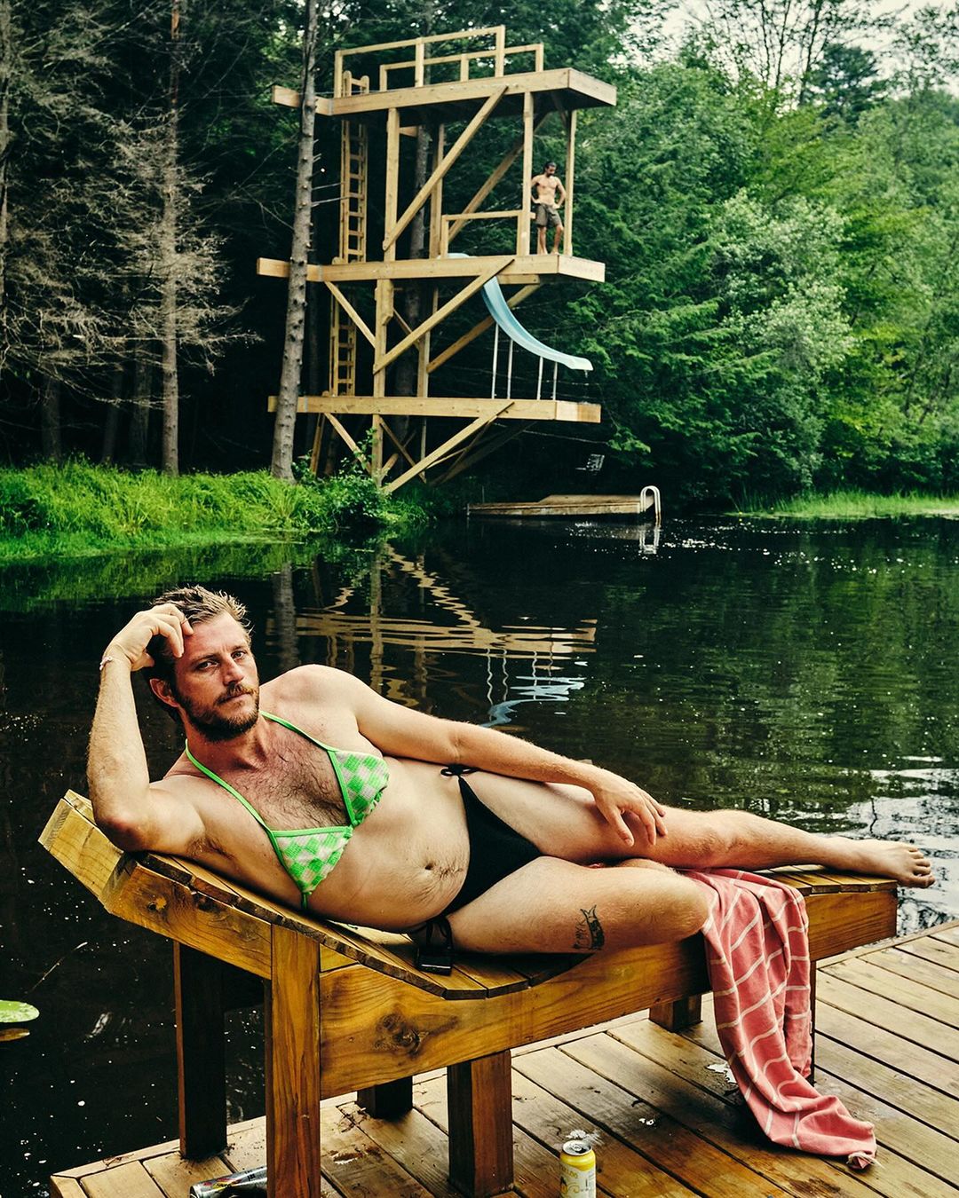 Man poses on a wooden lounge chair wearing a bikini near a pond with a wooden diving tower in the background on a summer day.