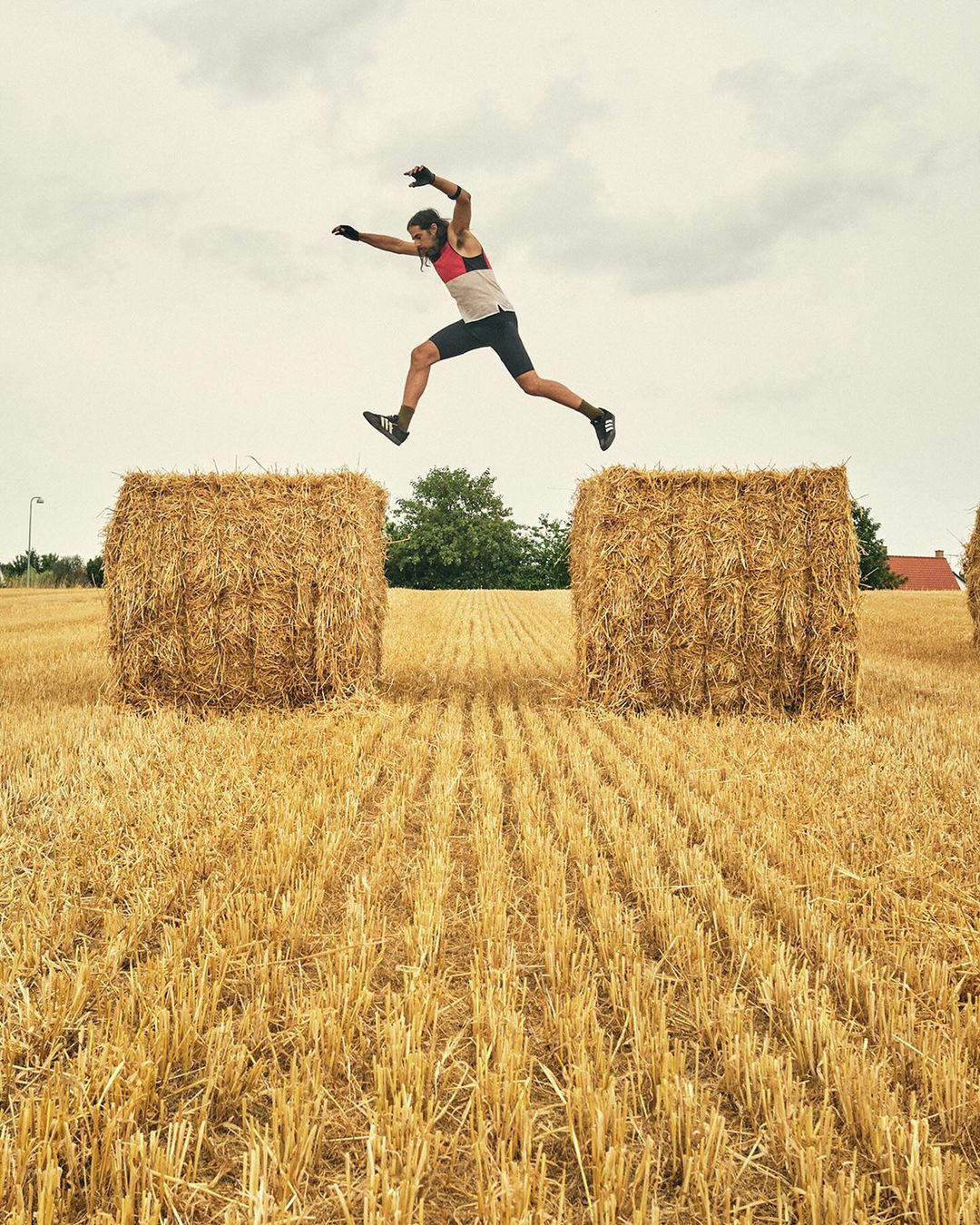 A man jumps over two large hay bales in a rural field under an overcast sky, showcasing athleticism and outdoor fitness.