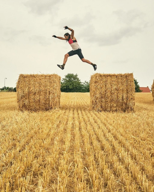 A man jumps over two large hay bales in a rural field under an overcast sky, showcasing athleticism and outdoor fitness.