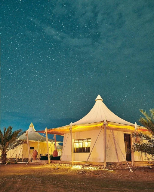 Two white canvas tents with wooden frames glow under a starry night sky in a desert landscape, creating a serene and inviting scene.