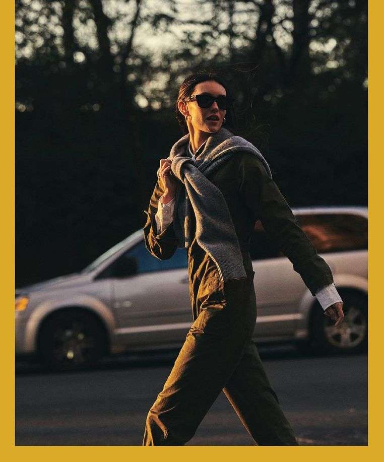 Woman in an olive green outfit and sunglasses walks on a road with a silver minivan visible in the background at dusk.