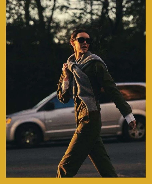 Woman in an olive green outfit and sunglasses walks on a road with a silver minivan visible in the background at dusk.