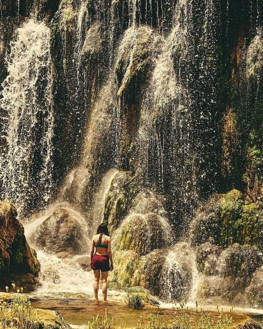Woman in swimwear standing in the river, looking at a tall waterfall surrounded by mossy rocks and plants in an outdoor setting.