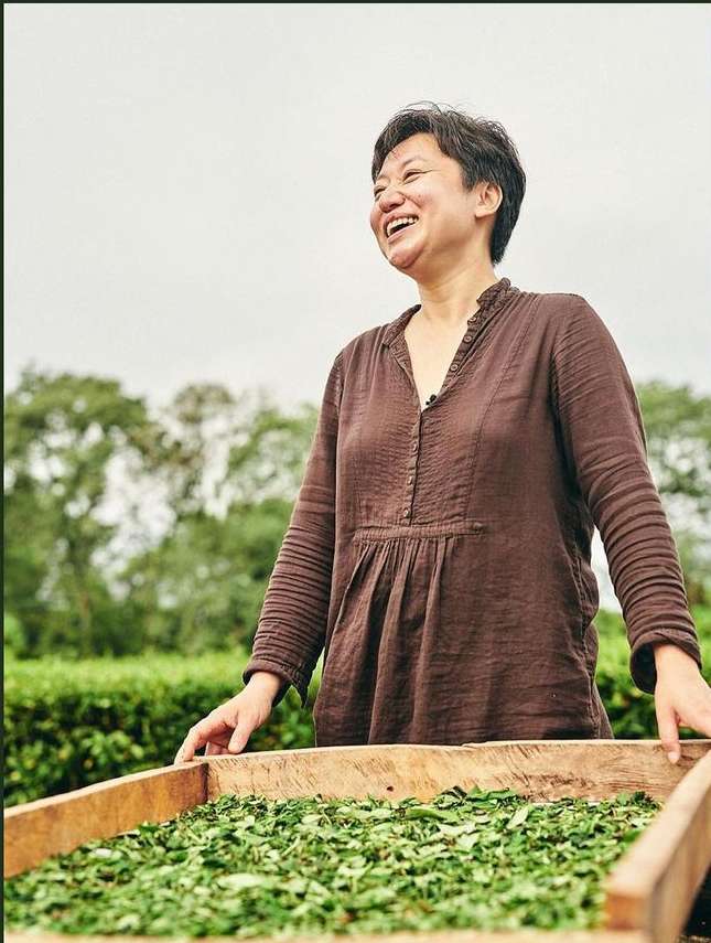 An adult woman smiles as she tends to a wooden tray of drying green tea leaves outdoors on a rural tea farm, trees in background.