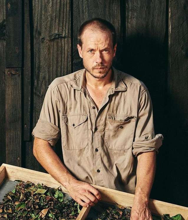 Portrait of a man in a tan shirt standing indoors holding a tray of dried leaves in a photo studio with a dark wooden background.