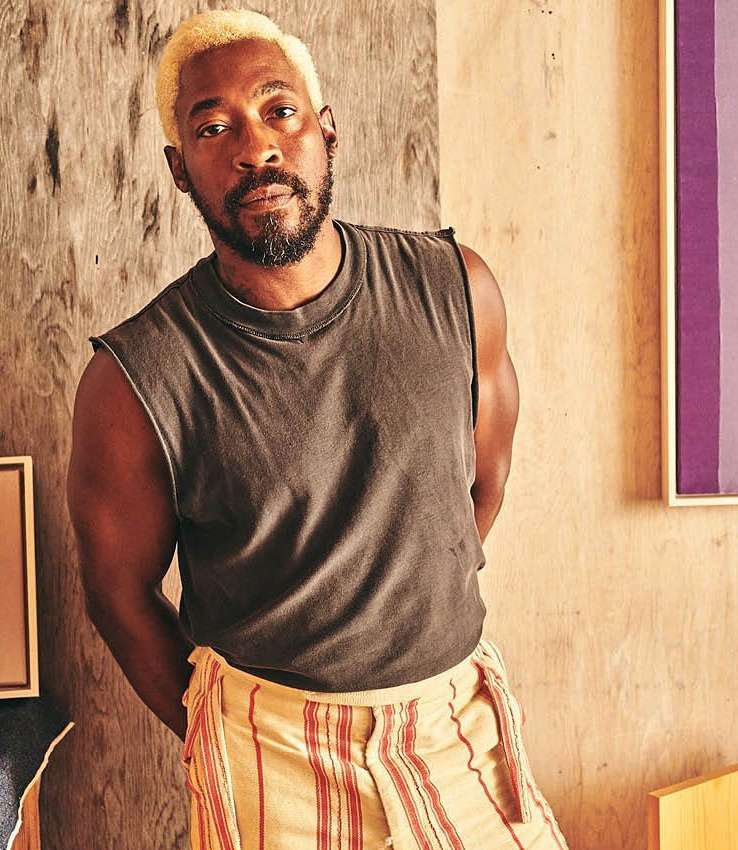 Adult male poses indoors wearing a grey sleeveless shirt and horizontally striped pants. He has short blonde hair and a beard.