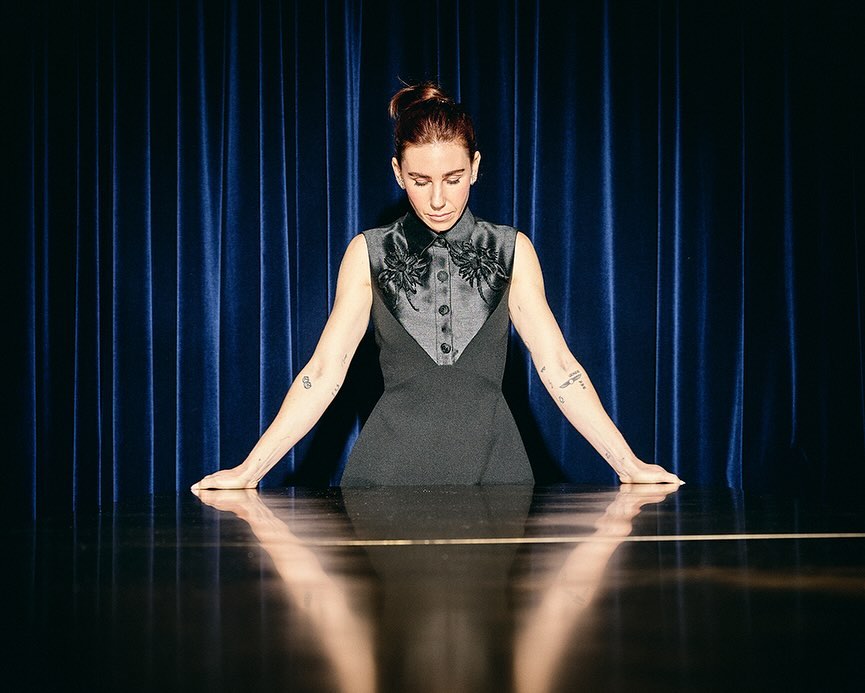 Portrait of a fair-skinned woman with an updo wearing a formal dark dress, standing behind a reflective table against a blue curtain backdrop.