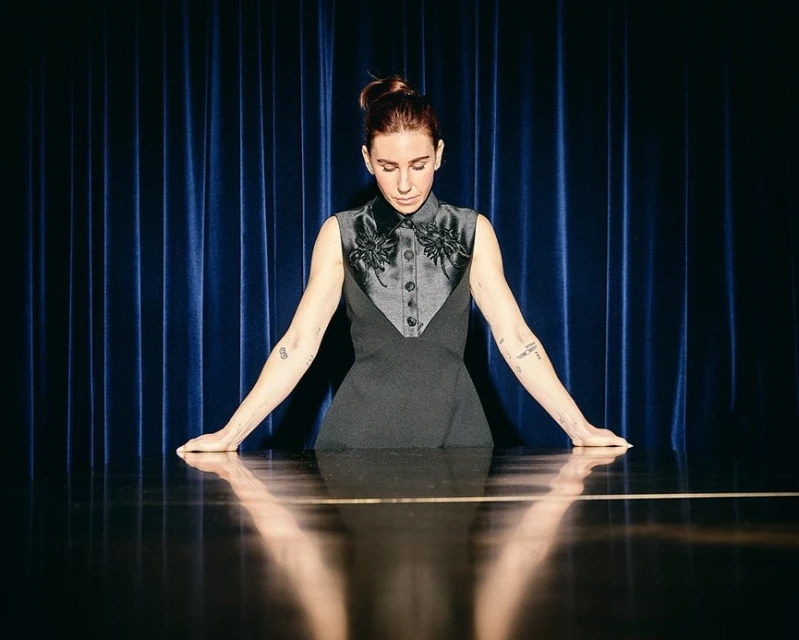 Portrait of a fair-skinned woman with an updo wearing a formal dark dress, standing behind a reflective table against a blue curtain backdrop.