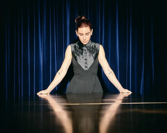 Portrait of a fair-skinned woman with an updo wearing a formal dark dress, standing behind a reflective table against a blue curtain backdrop.