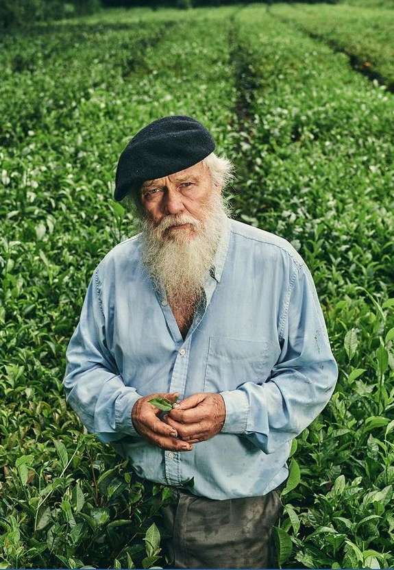 A man with a long white beard and a black beret stands in a lush green field, holding a leaf, creating a portrait of rural life and nature.