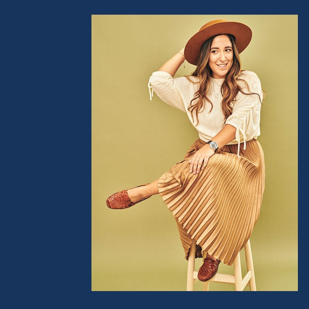 Woman in pleated skirt and hat sits on a stool in studio, smiling. She wears a peasant blouse and brown shoes.