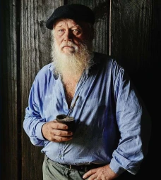 Elderly man with a white beard and black cap wearing a blue shirt and holding a gourd against a dark wooden wall.