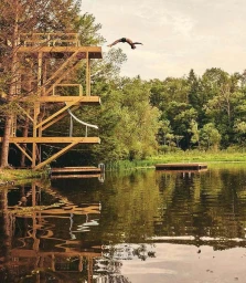 Man diving mid-air from a high wooden diving tower into a calm lake surrounded by lush green trees.