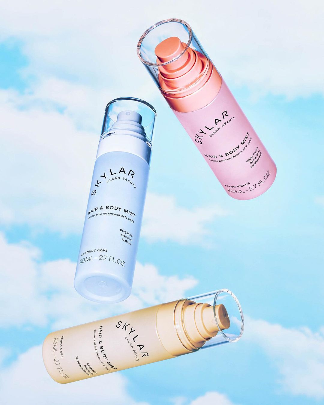 Three Skylar Hair & Body Mist bottles in light blue, pink, and beige float against a sky backdrop, showcasing beauty product photography.
