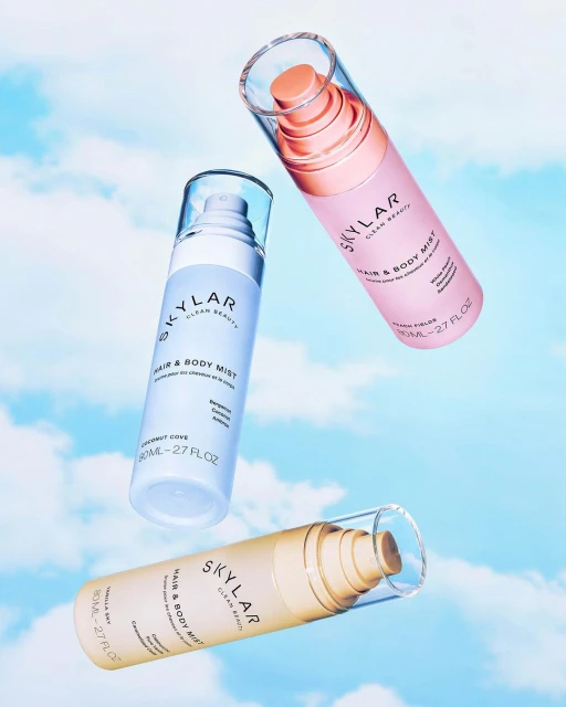 Three Skylar Hair & Body Mist bottles in light blue, pink, and beige float against a sky backdrop, showcasing beauty product photography.