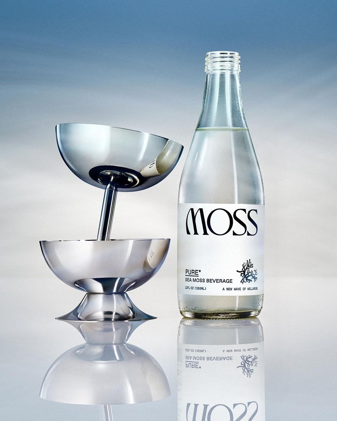 Elegant still life showcases a sleek metal cocktail tower beside a bottled beverage against a graduated blue backdrop with reflective surface.