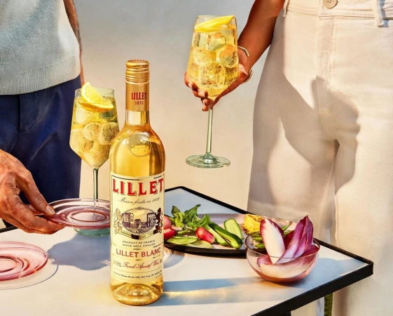 Lillet Blanc bottle with two people holding glasses of white wine with lemon slice, served with fresh vegetable snacks on a white table.