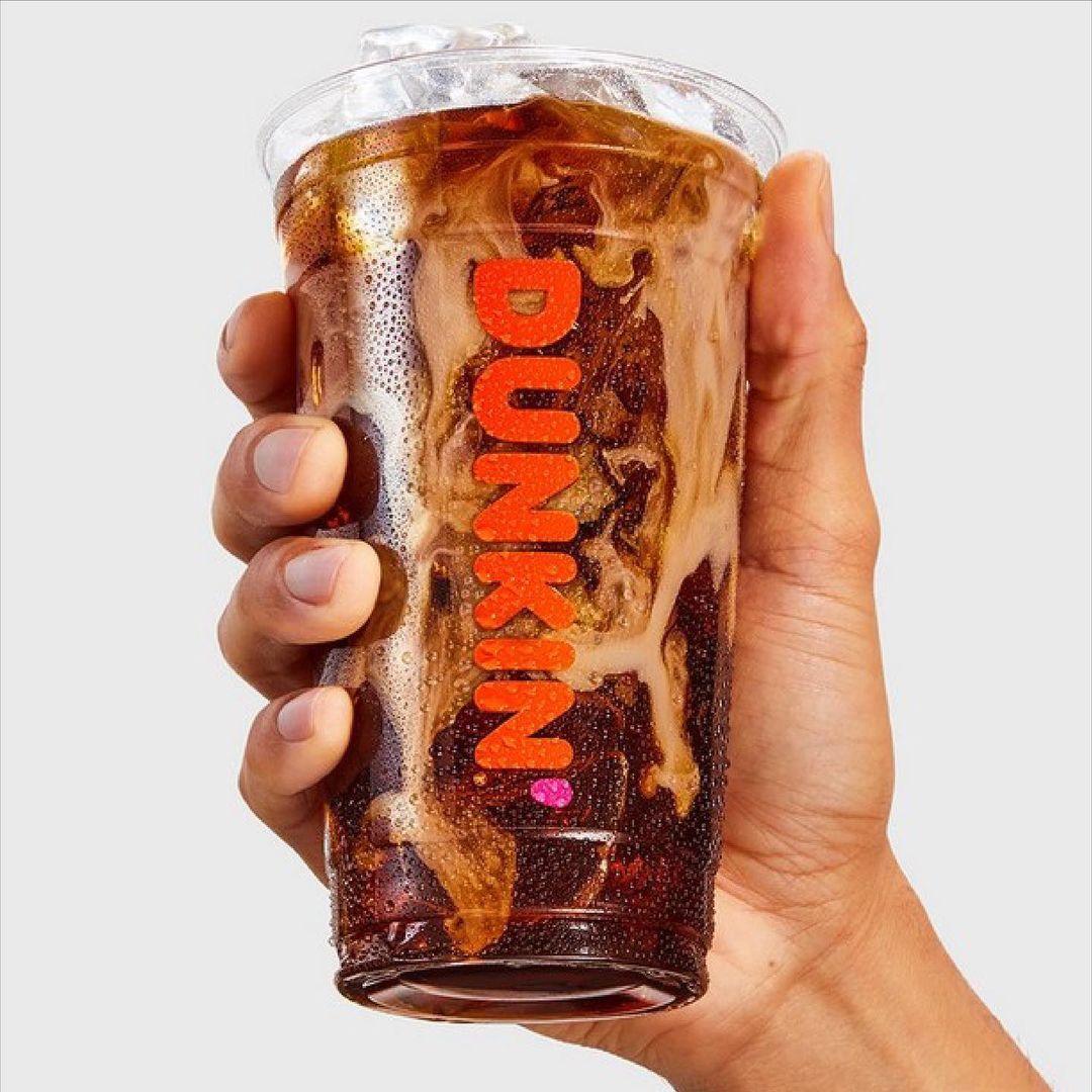 A hand holds a Dunkin' iced coffee in a clear plastic cup, showcasing the drink's creamy swirl and brand logo against a neutral background.