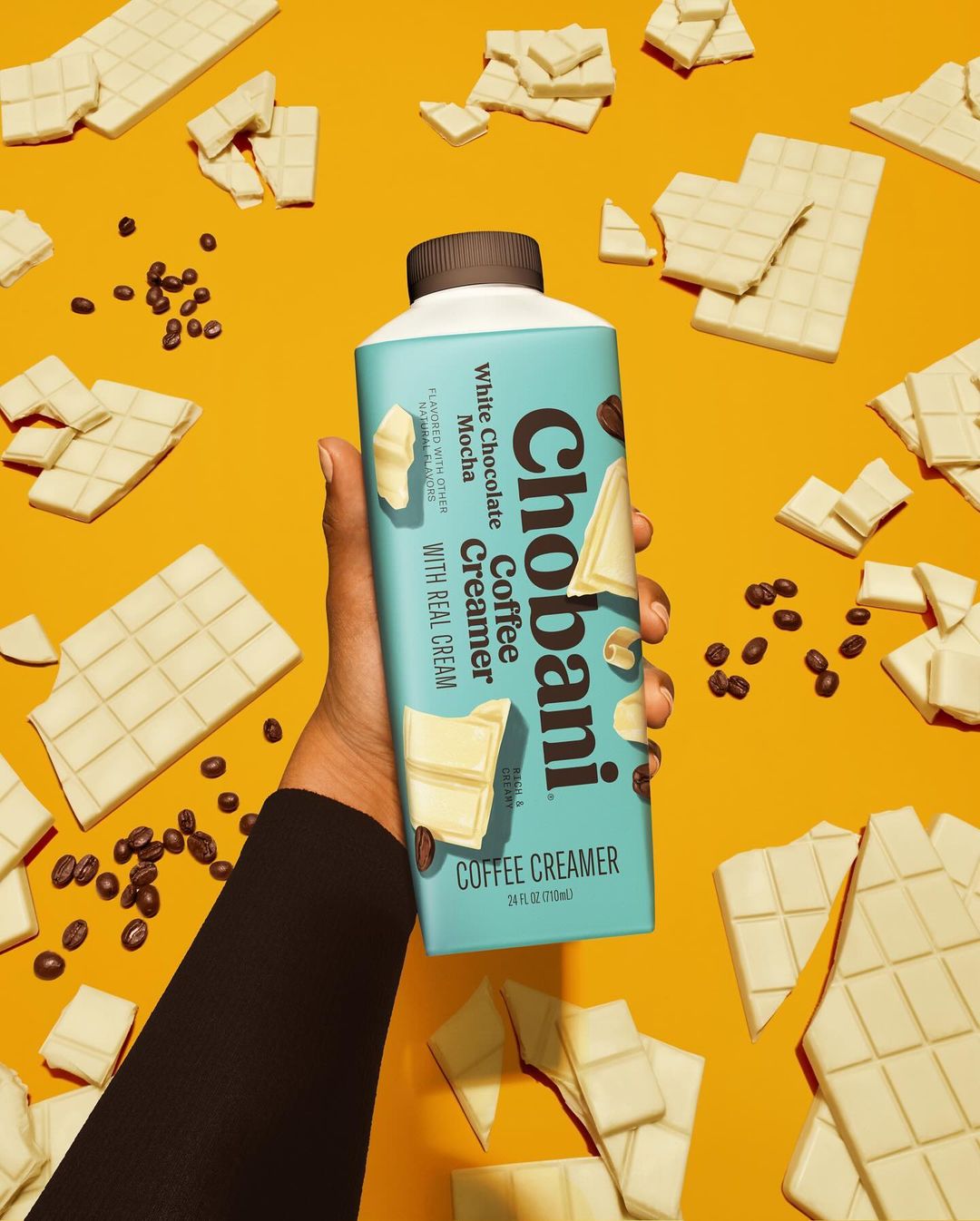 Hand holds Chobani White Chocolate Mocha Coffee Creamer with coffee beans and white chocolate squares on a vibrant yellow background.