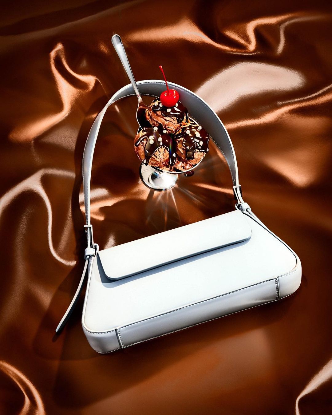 White purse draped over a chocolate ice cream sundae with a cherry on top, all against a brown satin background.