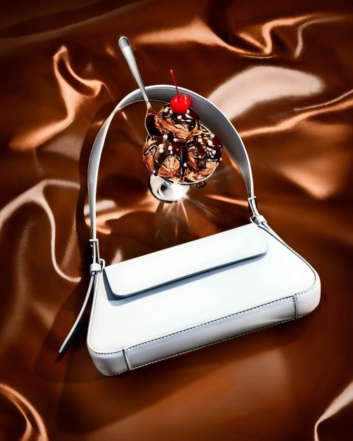 White purse draped over a chocolate ice cream sundae with a cherry on top, all against a brown satin background.