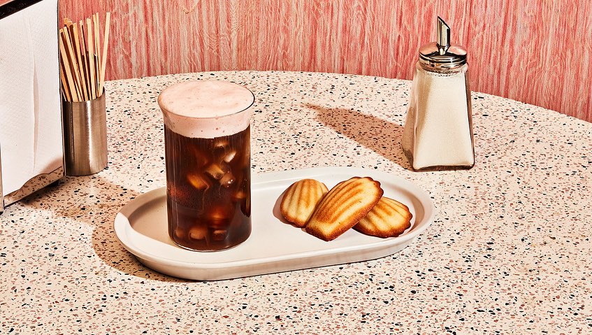 Iced coffee with milk foam accompanied by three madeleines on a white plate next to a sugar dispenser and napkin holder.
