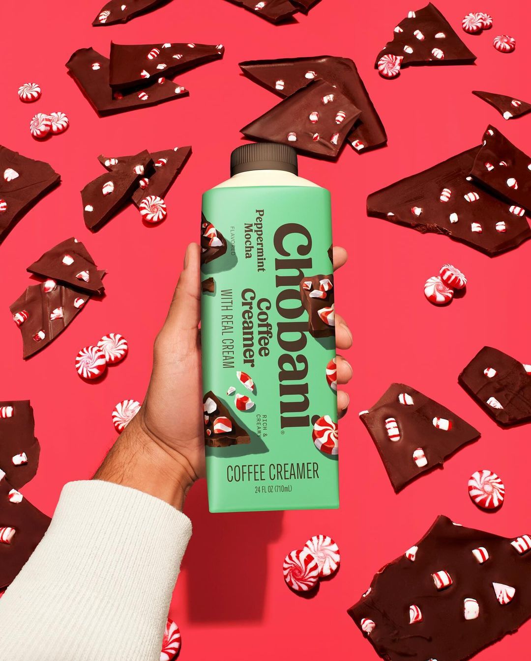 Person holding a carton of Chobani Peppermint Mocha Coffee Creamer surrounded by peppermint candies and dark chocolate pieces on a red backdrop.
