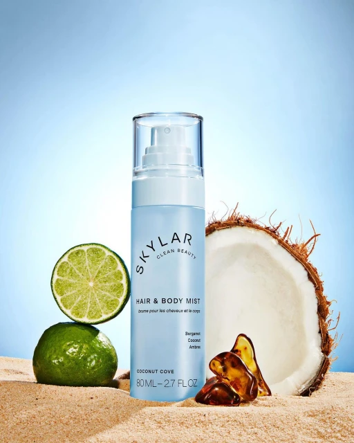 Skylar Clean Beauty hair and body mist displayed with coconut, lime, and amber on a sandy surface against a light blue gradient.